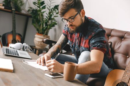 Young guitarist hipster at home with guitar writing notes drinking coffeeの写真素材