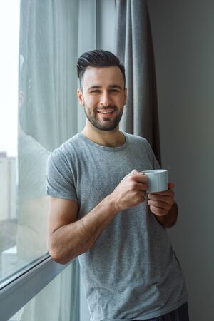 Bachelor man daily routine standing near the window single lifestyle concept drinking coffee smilingの写真素材