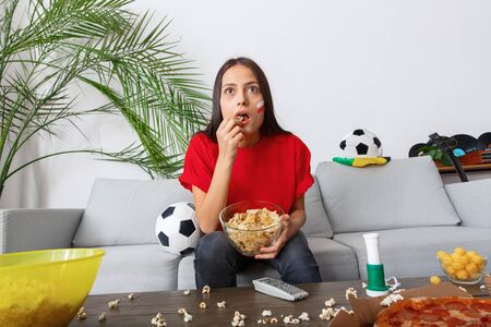 Young woman sport fan watching match in a red t-shirt eating chipsの写真素材