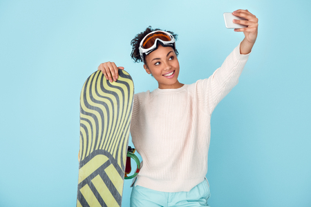 Young african woman isolated on blue wall studio winter sport snowboarding concept selfie photosの写真素材