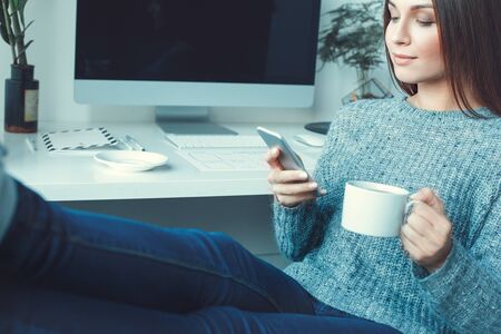 Young woman freelancer indoors home office concept casual style drinking tea browsing smartphoneの写真素材
