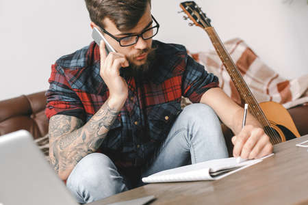 Young guitarist hipster at home with guitar sitting writing song phone call close-upの写真素材