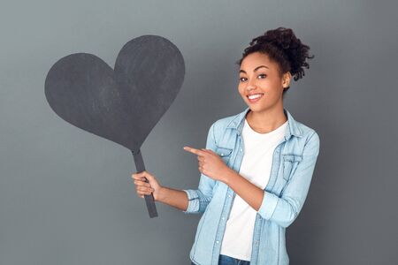 Young african woman isolated on grey wall studio casual daily lifestyle pointing at heart shapeの写真素材