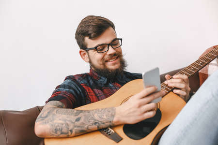 Young guitarist hipster at home holding guitar sitting browsing smartphone communicationの写真素材