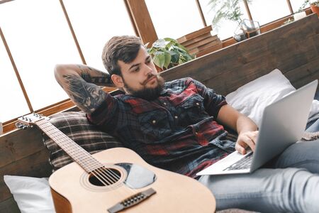 Young guitarist hipster at home with guitar in bedroom browsing internetの写真素材