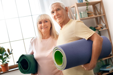 Senior couple exercise together at home health care holding yoga mats looking cameraの写真素材