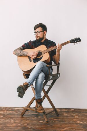 Young guitarist hipster at home with guitar on the chair full-body portraitの写真素材