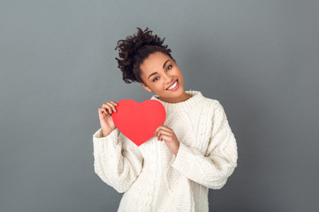 Young african woman isolated on grey wall studio winter concept holding heartの写真素材