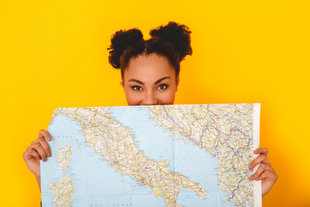 Young african woman isolated on yellow wall studio teen style traveler hiding behind mapの写真素材