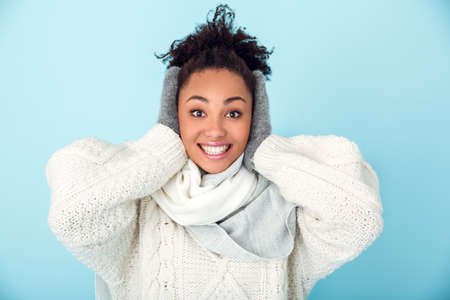 Young african woman isolated on blue wall studio winter concept cover earsの写真素材