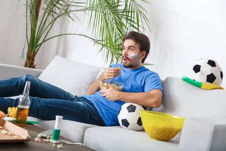 Young man sport fan watching match in a blue t-shirt eating chipsの写真素材