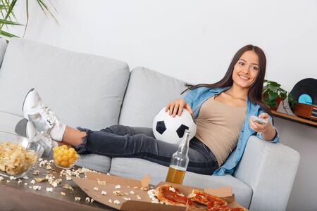 Young woman sport fan watching match changing volume with remote controllerの写真素材