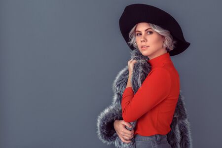 Young woman in a red blouse and fur coat isolated on grey wall taking of coatの写真素材