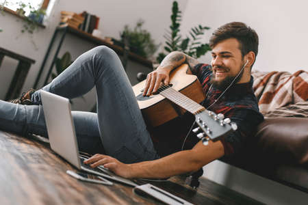 Young guitarist hipster at home sitting on the floor holding guitar earphones music browsing laptopの写真素材