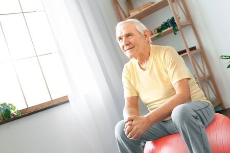 Senior man exercise at home sitting on exercise ball thinkingの写真素材