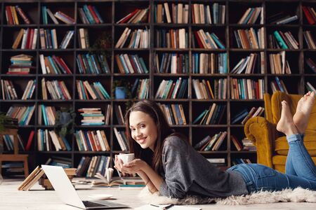 Young woman student in library at home lying drinking coffeeの写真素材