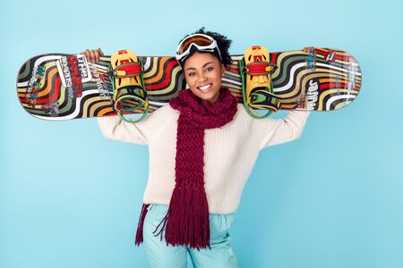 Young african woman isolated on blue wall studio winter sport snowboarding concept holding boardの写真素材
