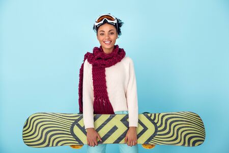 Young african woman isolated on blue wall studio winter sport snowboarding concept holding snowboardの写真素材