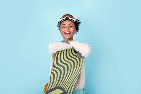 Young african woman isolated on blue wall studio winter sport snowboarding concept hugging boardの写真素材
