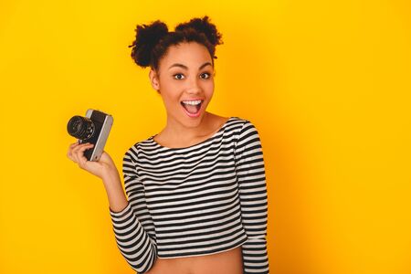 Young african woman isolated on yellow wall studio teen style holding cameraの写真素材