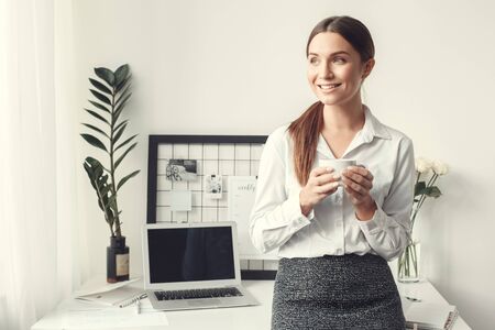 Young woman freelancer indoors home office concept formal style leaning on desk drinking teaの写真素材