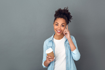 Young african woman isolated on grey wall studio casual daily lifestyle drinking cofee having phone callの写真素材