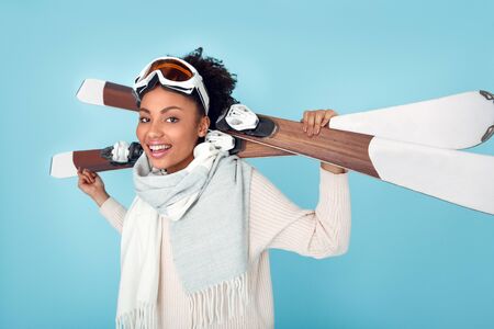 Young african woman isolated on blue wall studio winter sport skiing standing with skis side viewの写真素材