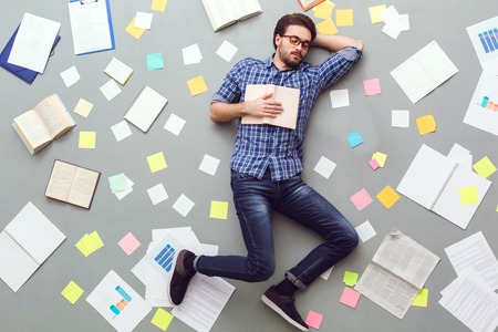 Young man isolated on grey background with papers and notes sleeping with bookの写真素材