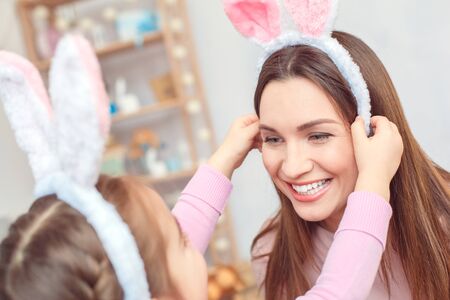 Mother and daughter in bunny ears easter celebration together at home sitting girl putting bazel on moms head close-upの写真素材