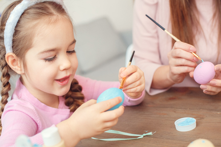 Mother and daughter easter celebration together at home sitting girl painting egg close-upの写真素材