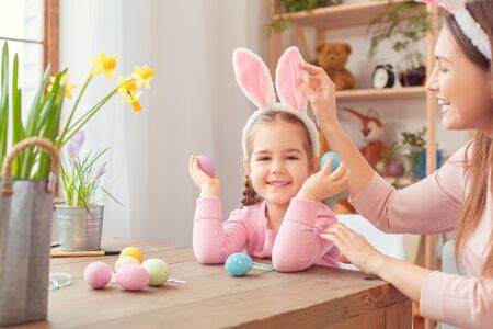 Mother and daughter in bunny ears easter celebration together at home sitting girl holding eggsの写真素材