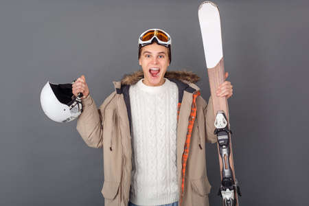 Young man studio isolated on grey winter style holding helmet and skis excitedの写真素材