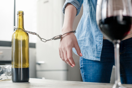 Young female alcoholic social problems standing near table fasten with handcuffs to wine bottle close-upの写真素材