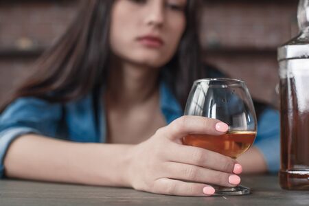 Young female alcoholic social problems sitting at table holding glass with whiskey close-up looking...