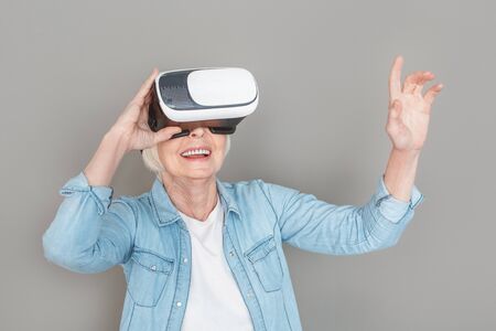 Aged female wearing jeans jacket studio isolated on grey background playing virtual reality smiling cheerful excitedの写真素材