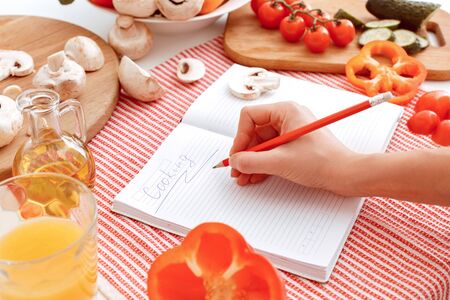 Hand writes a recipe. Many vegetables on the table and a notebook with an inscription cooking. Indoor, studio shoot, kitchen interior. Indoor, studio shoot, kitchen interiorの写真素材