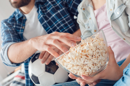 Bearded man and little girl at home family time sitting on sofa watching tv eating popcorn close-upの写真素材
