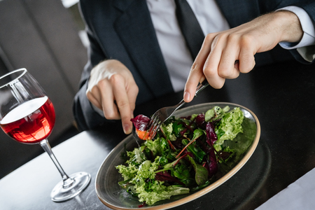 Businessman on business lunch at restaurant sitting at table eating fresh vegetable salad holding fork and knife drinking red wine close-upの写真素材