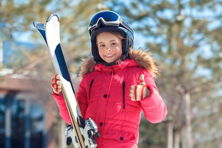 Winter vacation. Girl in goggles and helmet with skis standing outdoors smiling happy showing thumb up close-up blurred backgroundの写真素材