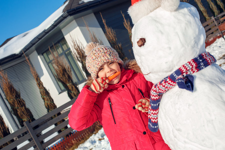 Winter vacation. Girl standing outdoors near house making snowman eating carrot nose smiling playful close-upの写真素材