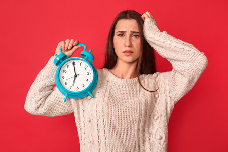 Late for Meeting. Young woman standing isolated on red with alarm clock touching head concernedの写真素材