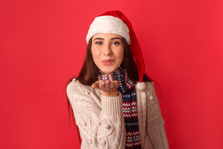 Celebration Concept. Young woman in scarf and santa hat standing isolated on red sending kiss to camera smiling playful close-upの写真素材