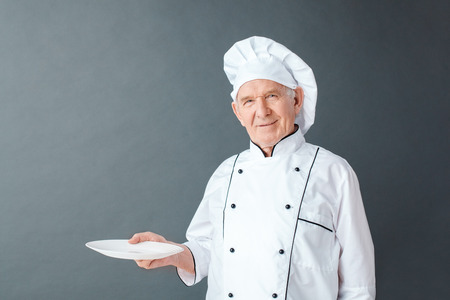 Senior chef studio standing isolated on gray with empty plate looking camera happyの写真素材