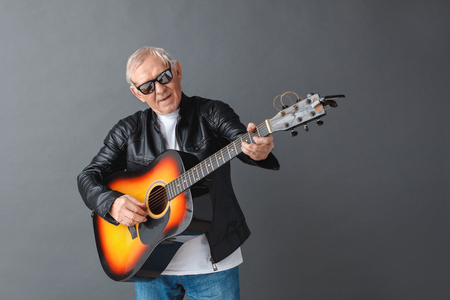 Senior man in leather jacket and sunglasses standing isolated on gray playing guitar singing joyfulの写真素材