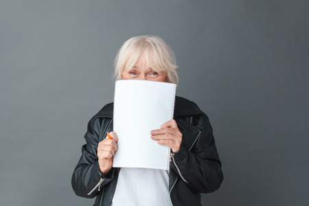 Senior woman in leather jacket studio standing isolated on gray covering face with notebook looking camera playful close-upの写真素材