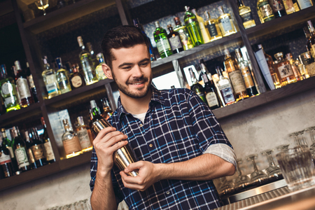 Young bartender standing at bar counter shaking shaker smiling thoughtfulの写真素材