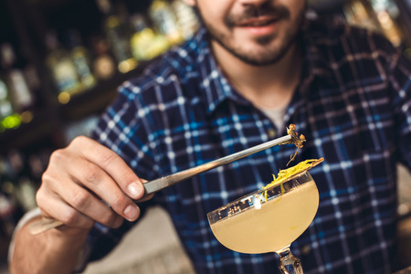 Young bartender standing at bar counter decorating cocktail with herb smiling close-upの写真素材
