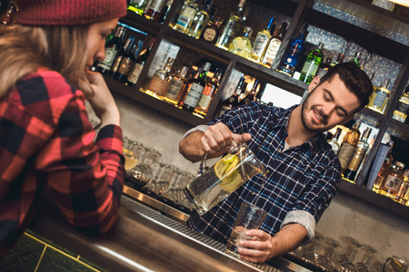 Young bartender standing at bar counter serving client pouring glass of water smilingの写真素材