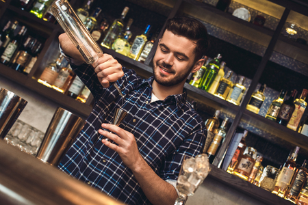 Young bartender standing at bar counter pouring alcohol into jigger happyの写真素材