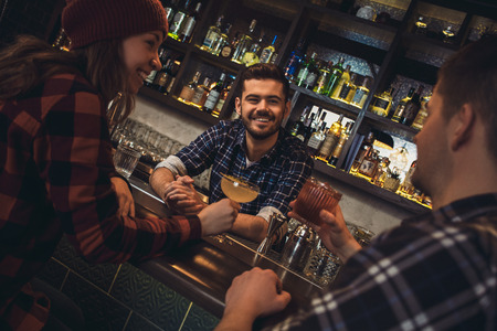 Young bartender standing at bar counter with visitors drinking alcohol talking joyfulの写真素材